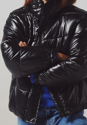 Personne croisant les bras portant une doudoune noire brillante, un pull bleu et un jean bleu sur un fond neutre.