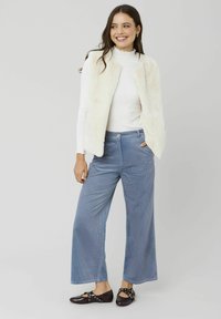 Gilet en fausse fourrure crème, haut à manches longues blanc, pantalon en velours côtelé large bleu clair et chaussures noires avec des accents argentés.