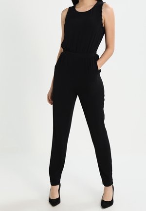 Vrouw draagt een zwarte mouwloze jumpsuit met taps toelopende pijpen en zwarte hoge hakken tegen een witte achtergrond.