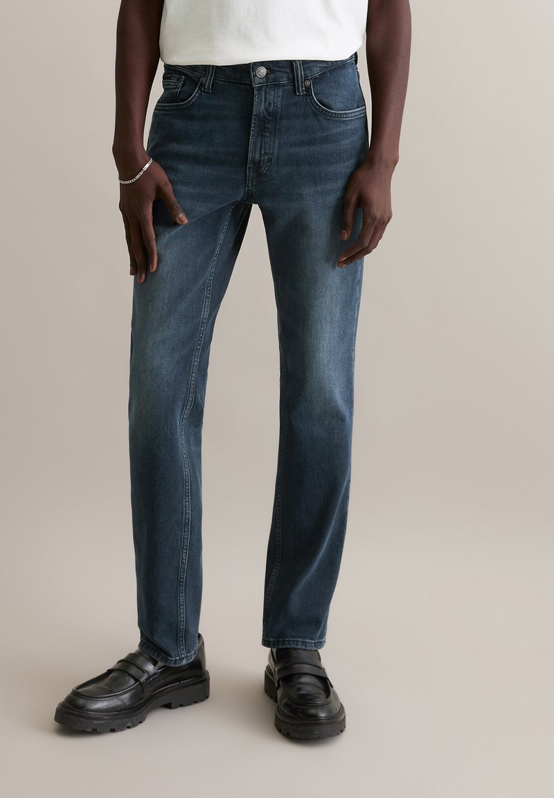 Jean en denim bleu foncé de coupe droite, cinq poches et fermeture à bouton. Porté avec des mocassins noirs. Texture lisse et délavage subtil.