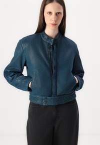 Chaqueta de mezclilla en azul intenso con un cuello alto, cremallera frontal y dos botones visibles en la cintura; cuenta con hombros caídos y un corte corto.