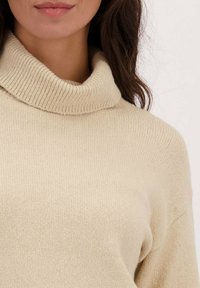 Beige coltrui gemaakt van zachte, ribbelstof. Heeft een hoge, gevouwen kraag en een ontspannen pasvorm. Lichtjes gestructureerd oppervlak.