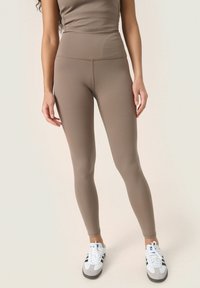 Barna magas derekú leggings rugalmas anyagból, sima felülettel és testhez simuló kialakítással, fehér sportcipővel párosítva.