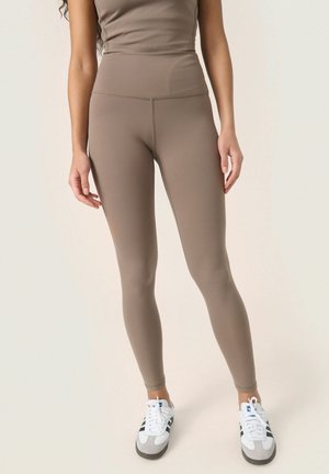 Bruine high-waisted leggings van stretchstof, met een gladde textuur en een getailleerd ontwerp, gecombineerd met witte sportschoenen.