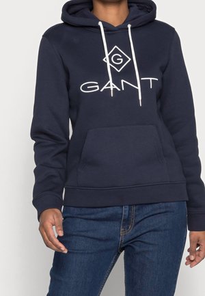Personne portant un sweat à capuche GANT bleu marine avec des cordons blancs et une poche avant, associé à un jean bleu, debout devant un fond clair.