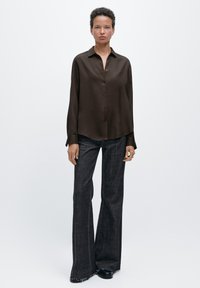 Camicia marrone a maniche lunghe con bottoni e consistenza morbida; abbinata a pantaloni svasati scuri e scarpe nere, che mostrano una vestibilità comoda e un design minimalista.