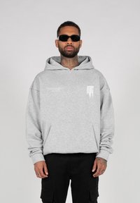 Sudadera gris de gran tamaño hecha de un tejido suave, con un bolsillo frontal y un logo blanco en el lado izquierdo del pecho. Combinada con pantalones cargo negros.