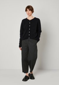 Tumši zili džemperis, izgatavots no mīkstas, strukturētas auduma, ar apaļu kakla izgriezumu un sešām baltajām pogām, kombinēts ar brīviem melniem biksēm un melnām kurpēm.