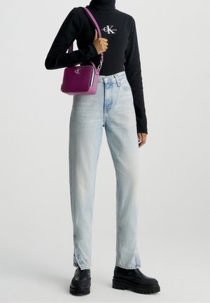 Persona che indossa jeans blu chiaro con orli a spacco, stivali neri con plateau, maglione nero con colletto alto e logo bianco, mentre porta una piccola borsa a tracolla viola.