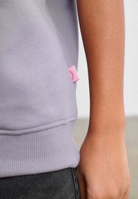 Levandulová mikina s žebrovaným lemem a růžovou štítkem na boku. Detailní pohled na látku a ruku opírající se o ni.