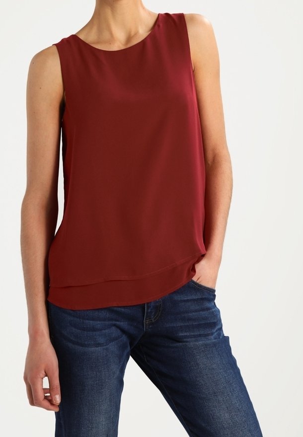 Femme portant un chemisier rouge foncé sans manches et un jean bleu, avec une main dans la poche, sur un fond blanc uni.