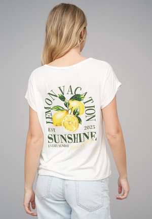 Pitkähiuksinen vaaleatukkainen nainen, joka pukeutunut valkoiseen t-paitaan, jonka selässä on sitruunoita ja vihreä teksti "Lemon Vacation Sunshine".