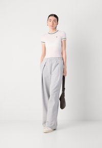Hellrosa, enganliegendes T-Shirt mit grünen Akzenten, kombiniert mit lockeren grauen Sweatpants. Das Model hält eine braune Umhängetasche und trägt weiße Schuhe.