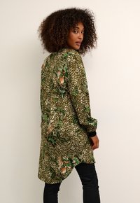 Chemise longue à coupe ample en imprimé floral vert et beige. Caractéristiques d'un ourlet incurvé, de manches longues avec des poignets contrastés, et d'un tissu doux et texturé.