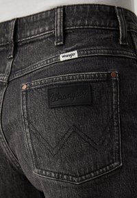 Czarne dżinsy z denimu posiadają na tylnej kieszeni naszywkę z logo Wrangler, miedziane nity oraz zygzakowate przeszycia na kieszeniach.