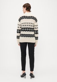Gestrickter cremfarbener Pullover mit schwarzen geometrischen Mustern, Rollkragen und geripptem Saum. Kombiniert mit schwarzen, eng anliegenden Hosen und schwarzen High Heels.