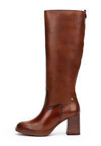 Botte en cuir marron, haute, avec un talon bloc, des coutures décoratives et un petit logo métallique sur le côté, présentée sur un fond blanc.