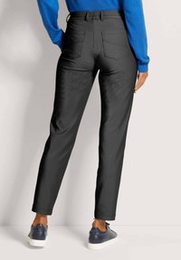 Persoon in donkergrijze high-waisted broek, blauwe langslavende top en zwarte leren sneakers, staande tegen een lichte achtergrond.