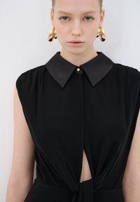 Robe noire avec un col arrondi, détail de bouton et ouverture en forme de clé à la taille. Présente des épaules froncées pour une texture supplémentaire. Boucles d'oreilles en or en forme de cercle.