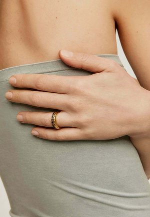 Anillo dorado y negro con un diseño texturizado, mostrado sobre una mano que descansa contra un fondo de tela gris claro.