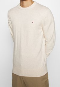 Beige stickad tröja med rund halsringning och långa ärmar. Har en liten flerfärgad logotyp på bröstet. Mjuk yta.