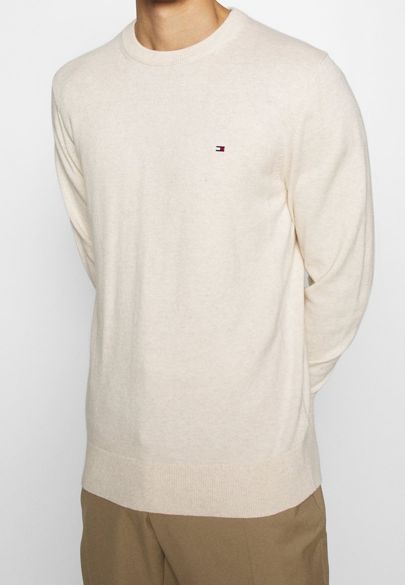 Beige stickad tröja med rund halsringning och långa ärmar. Har en liten flerfärgad logotyp på bröstet. Mjuk yta.