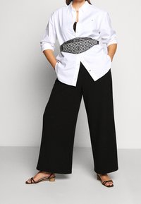 Donna che indossa una camicia bianca con bottoni, pantaloni larghi neri, sandali neri con cinghie e una cintura a tracolla con motivo in vita.