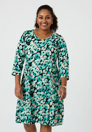 Smilende kvinde iført en knælængde kjole med et abstrakt blomsterprint i farverne grønblå, sort og beige, stående foran en ensfarvet baggrund.