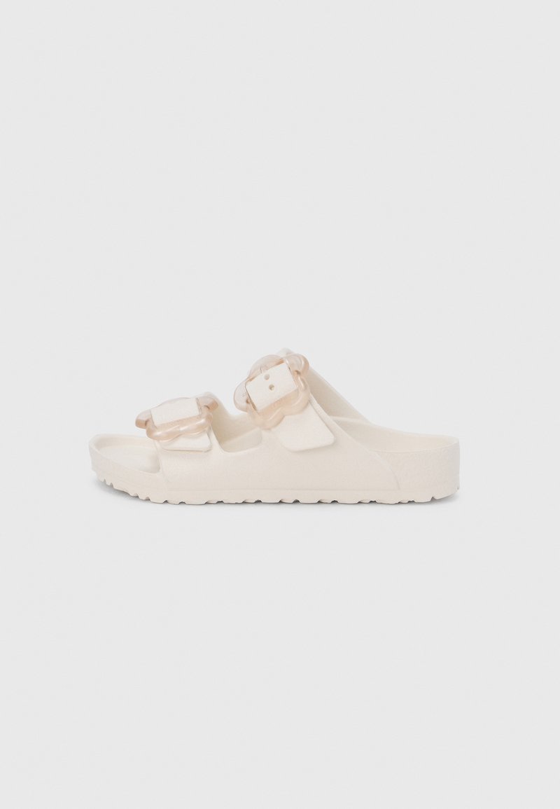 Λευκά σανδάλια με λουρί, κατασκευασμένα από μαλακό υλικό. Διαθέτουν δύο ρυθμιζόμενες διαφανείς αγκράφες με υφή στο λουρί και ανθεκτική σόλα.