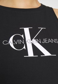 Czarny top bez rękawów z dużym białym logo "CK" oraz tekstem "CALVIN KLEIN JEANS" w nowoczesnej czcionce. Materiał wydaje się gładki i przypominający bawełnę.