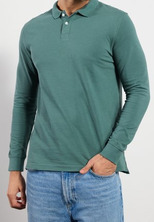 Homme portant un polo vert à manches longues avec deux boutons, associé à un jean bleu clair, main dans la poche, sur un fond uni.