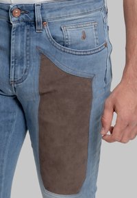 Jeans azzurri chiaro con una grande toppa in camoscio marrone scuro sulla coscia, cuciture marroni e un dettaglio con tasca frontale.