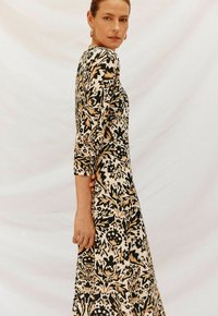 Abito lungo con stampa floreale nera, beige e dorata. Presenta maniche lunghe e una silhouette aderente. Il tessuto sembra texturizzato e leggero.