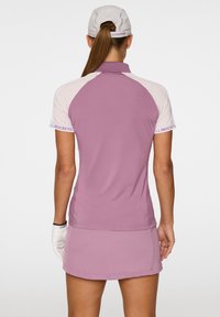 Maglietta polo viola con maniche rosa chiaro, dotata di colletto e accenti di logo visibili. Accompagnata da una gonna abbinata e guanti.