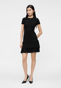 Μαύρο φόρεμα με κοντά μανίκια, εφαρμοστό πάνω μέρος και φούστα με στρώσεις βολάν. Κατασκευασμένο από υφασμάτινη ύλη με υφή, που διαθέτει ομαλή επιφάνεια και ρέουσα σιλουέτα.