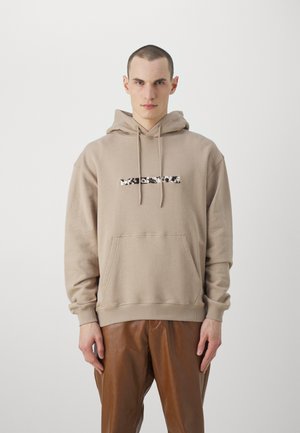 Beige hoodie met een voorzak en een trekkoord capuchon. Heeft een grafisch tekstontwerp in zwart en wit over de borst.