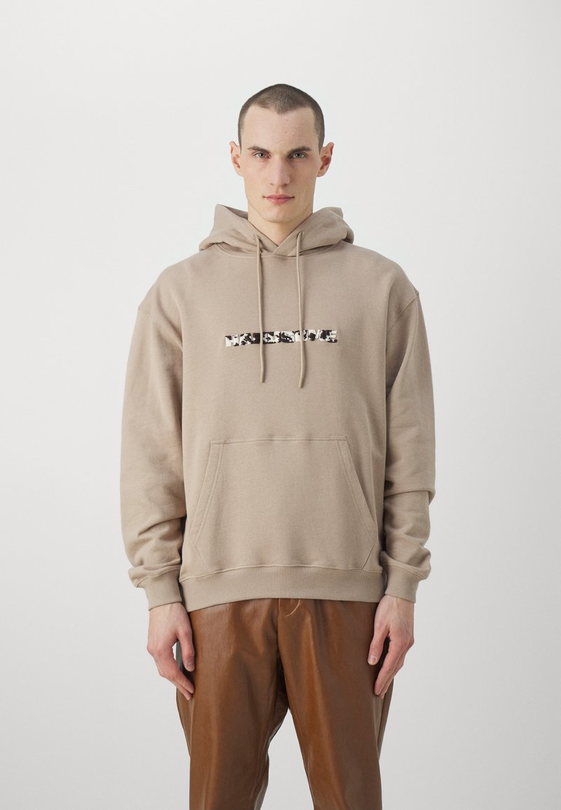 Sudadera beige con un bolsillo frontal tipo bolsa y capucha con cordón. Presenta un diseño gráfico de texto en blanco y negro en el pecho.