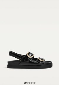 Sorte patentlæder sandaler med tre justerbare remme, hardware i guldfarve og en tekstureret sort sål. Bred pasform.