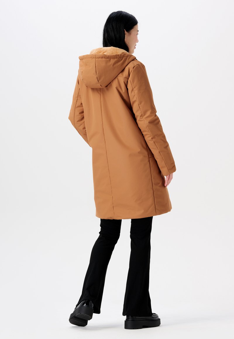 Langer brauner Puffercoat mit Kapuze, ausgestattet mit einem weichen Innenfutter und seitlichen Taschen. Tragend zu schwarzen, weit geschnittenen Hosen und klobigen schwarzen Schuhen.
