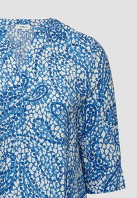 Lichte, blauw gestippelde blouse met een V-hals en driekwart mouwen, met abstracte vormen in verschillende tinten blauw en wit.