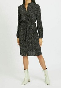 Žena v černých šatech ke kolenům s dlouhým rukávem a bílými vzory, s upnutým pasem, v kombinaci s bílými kotníkovými botami.