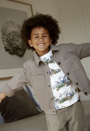 Tan knoopjasje over een witte blouse met patronen van bouwvoertuigen, gecombineerd met bijpassende broek, gemaakt van textuurstof en met een ontspannen pasvorm.