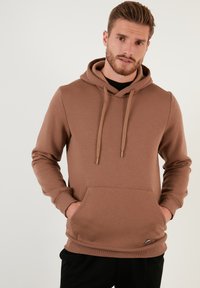 Sudadera marrón de tela suave, con un bolsillo tipo canguro, capucha ajustable con cordón y puños acanalados. Diseño simple, corte relajado.