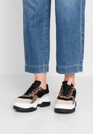 Osoba ubrana w niebieskie dżinsy i masywne sneakersy z czarnymi, beżowymi i panterkowymi detalami, stojąca na białej podłodze.