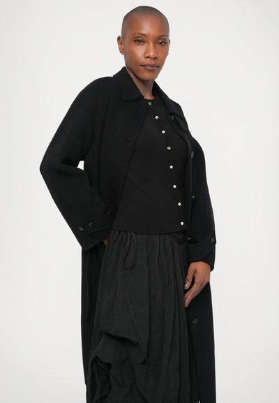 Largo abrigo negro con textura suave, mangas anchas y cierre de botones, superpuesto a una blusa negra con botones decorativos y una falda plisada.