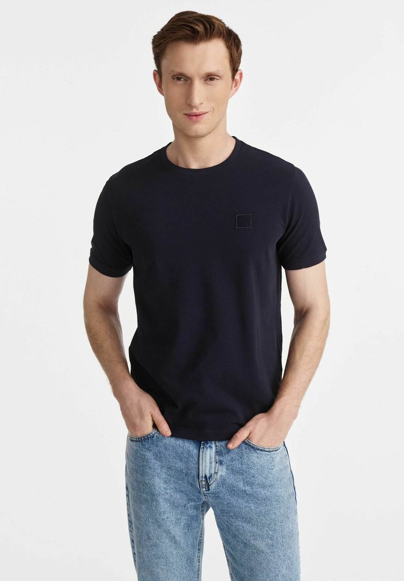 Granatowa koszulka z krótkim rękawem z bawełny, okrągły dekolt i małe prostokątne logo na piersi. W zestawie z jasnoniebieskimi dżinsami.