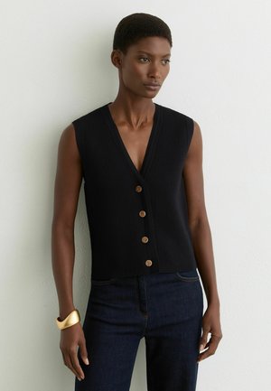 Kvinde med kort hår iført sort ærmeløs knappet vest og mørke jeans, stående op ad en lys, tekstureret væg, iført gyldt armbånd.