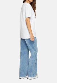 Witte katoenen t-shirt met korte mouwen, gecombineerd met lichtblauwe wijde jeans. Model draagt witte sneakers. Zijaanzicht.