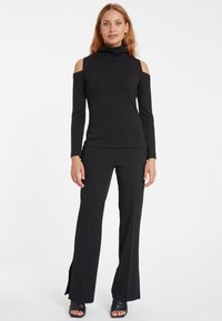Schwarzes Cold-Shoulder-Top mit hohen Ausschnitt und langen Ärmeln, kombiniert mit schwarzen Schlaghosen mit seitlichen Schlitzen. Glatter, dehnbarer Stoff.