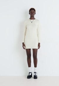 Robe en tricot crème à manches longues avec une silhouette ajustée. Le modèle porte des chaussures à plateforme noires et des chaussettes blanches. Encolure ronde simple, texture sans couture.